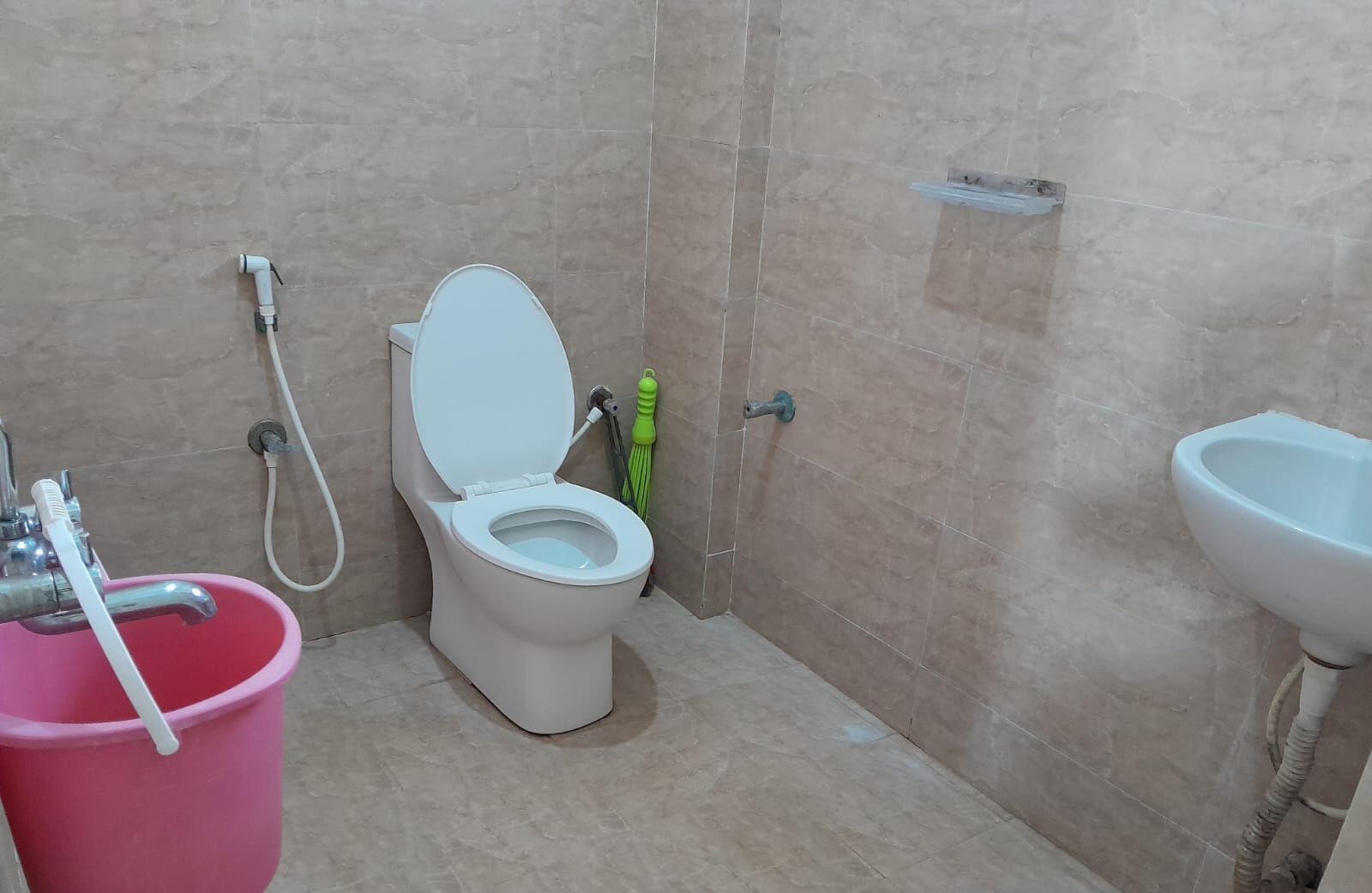 A bathroom with a toilet, a pink bucket, a green brush, and a white sink.