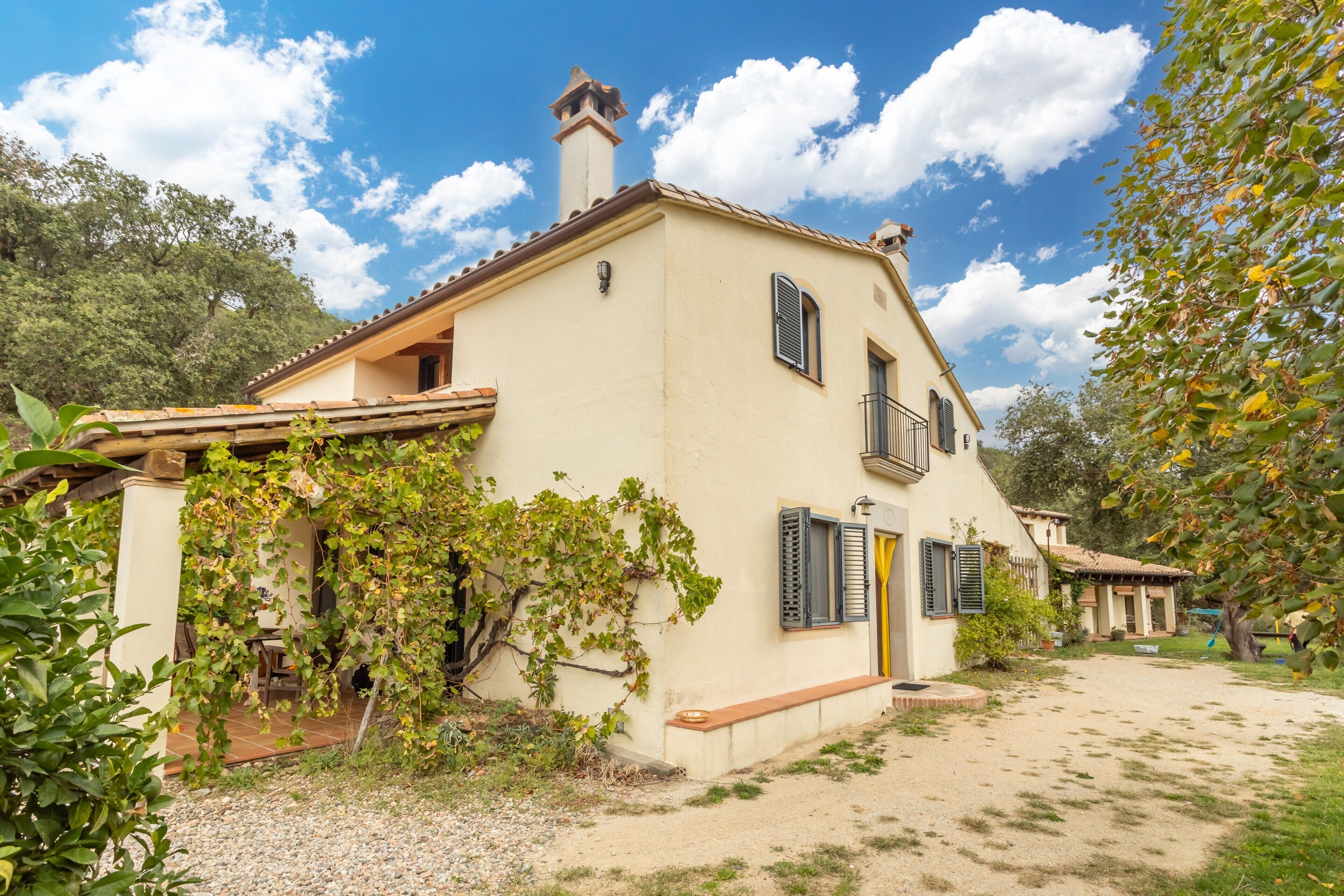 Una casa de dos pisos con un balcón, un jardín con un banco y un cielo despejado.