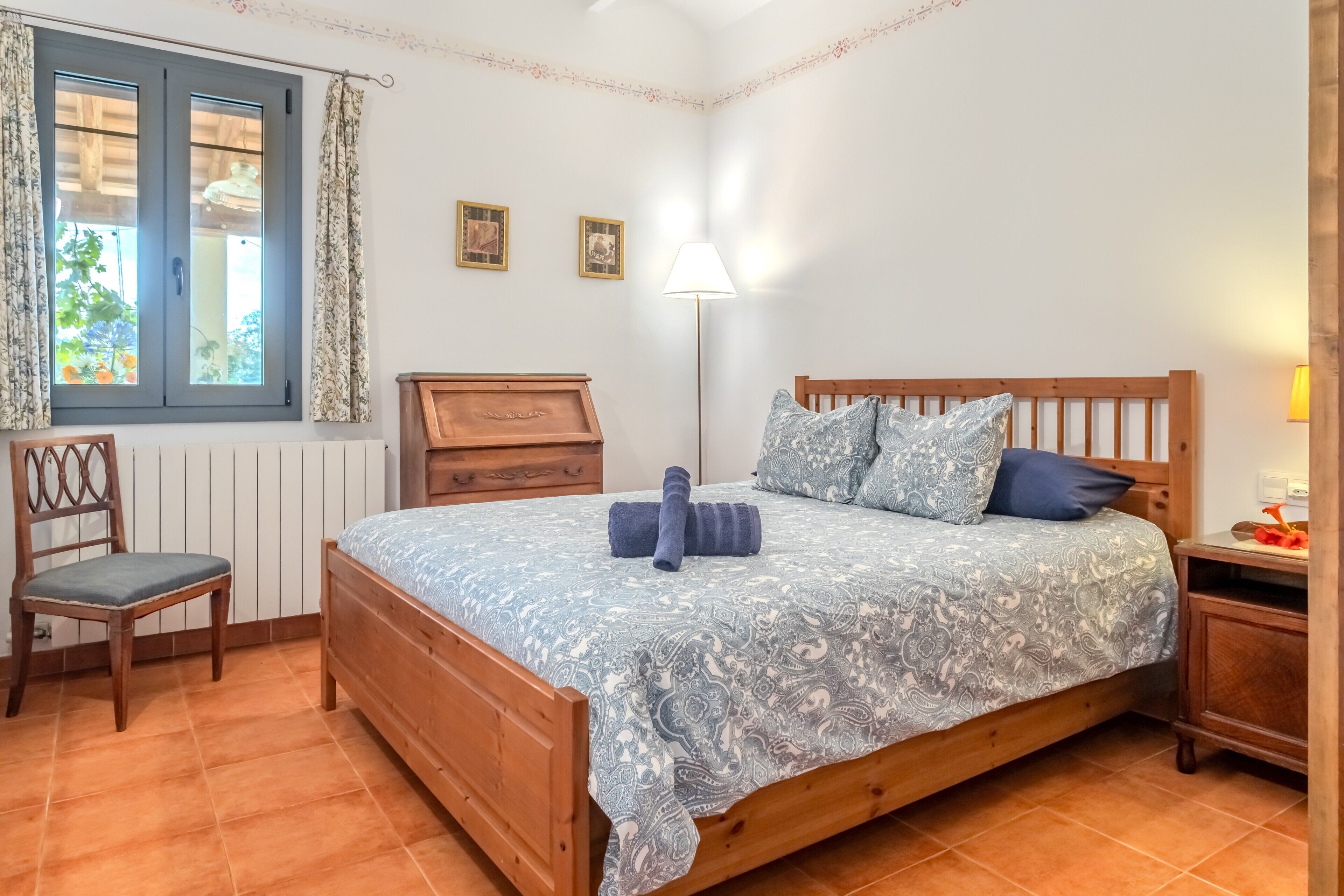 Ein Schlafzimmer mit einem hölzernen Bett, einem Sessel, einem Nachttisch, einer Lampe und einem Fenster mit Vorhängen.