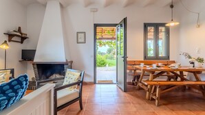 A cozy living room with a fireplace, a dining area with wooden benches, and a view through an open door to an outdoor garden. - Rural House 'Vinyoles 1' with Mountain View, Shared Pool and Wi-Fi (Quart)
