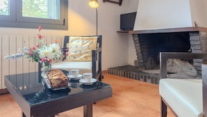 A cozy room with a fireplace, a table with a loaf of bread and cups, a chair with a floral cushion, and a lamp. - Rural House 'Vinyoles 1' with Mountain View, Shared Pool and Wi-Fi (Quart)