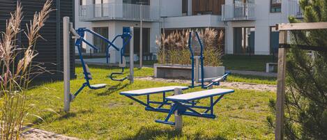 An outdoor gym area with exercise equipment, picnic tables, and a building with balconies in the background.