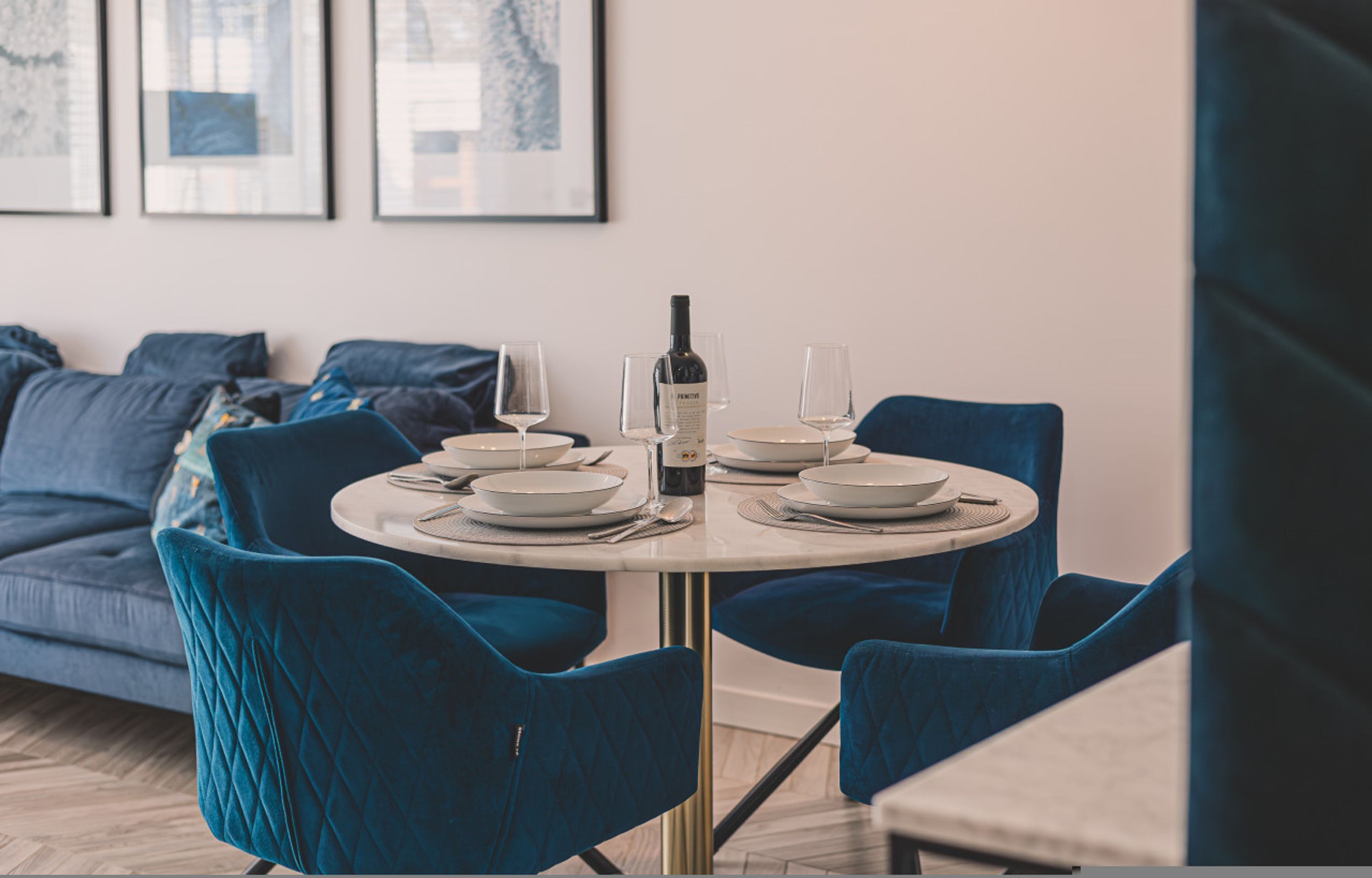 A dining area with a round table set for four, blue velvet chairs, a wine bottle, and white plates.