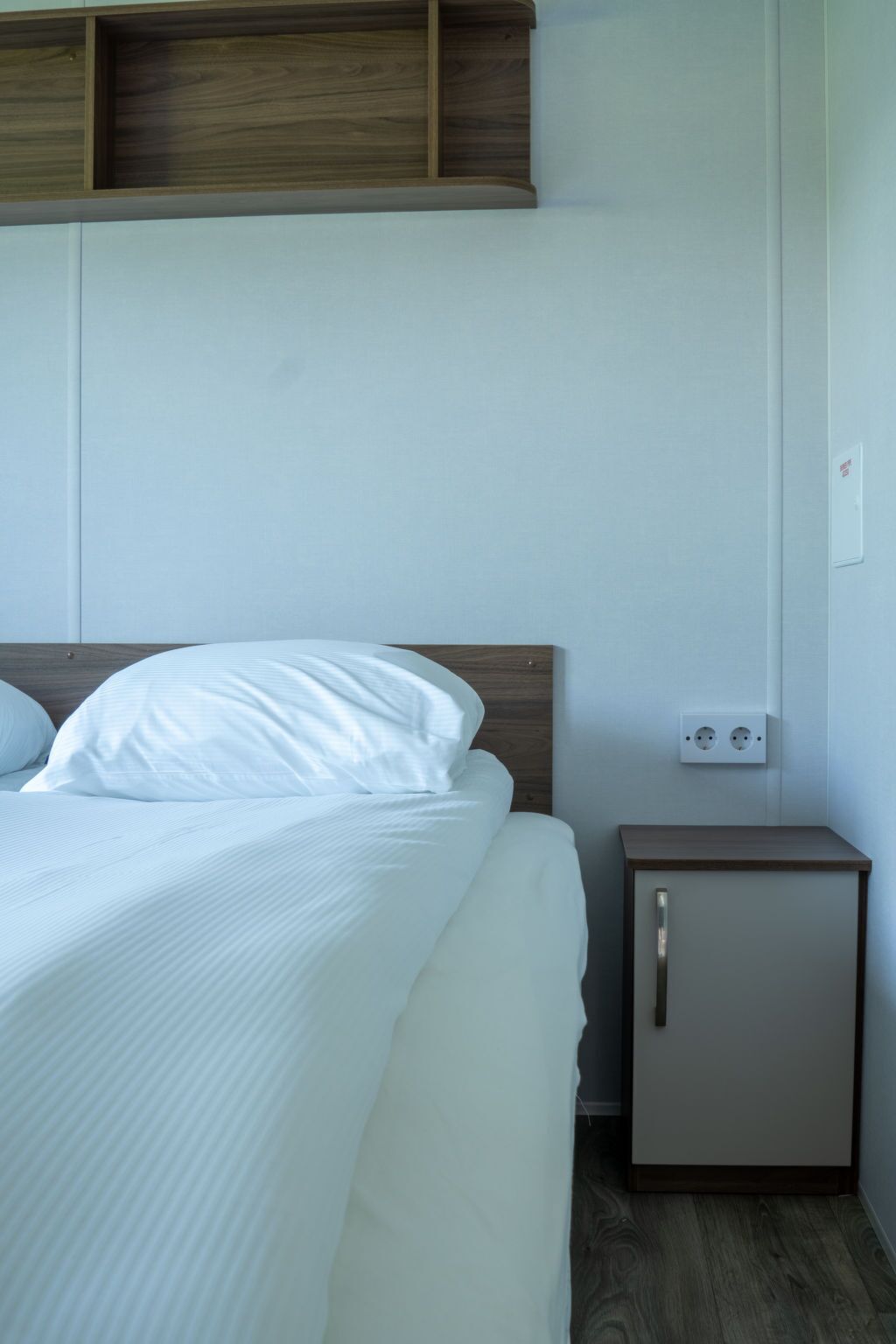 A neatly made bed with white linens, a wooden headboard, a small bedside table with a drawer, and a wall socket.