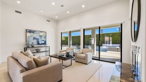 A spacious living room with a sofa, coffee table, and chairs, large windows with a view of a pool and greenery, and a modern art piece on the wall.