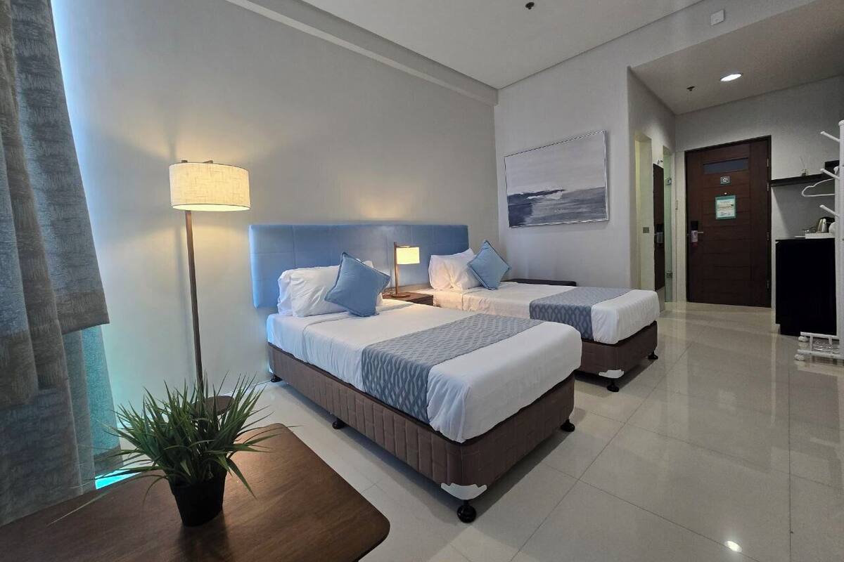A hotel room with two beds, a nightstand, a lamp, a plant, and a view of the sea.