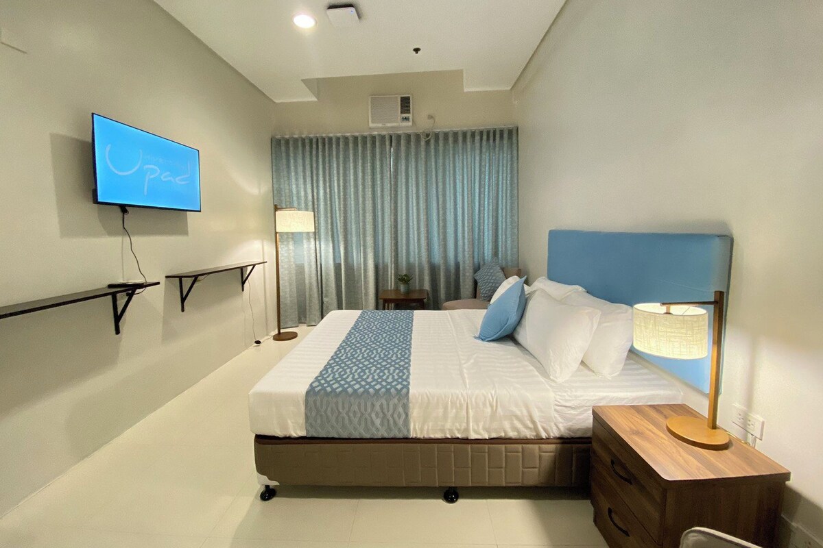 A modern hotel room with a bed, bedside tables, a TV, and a window with curtains.