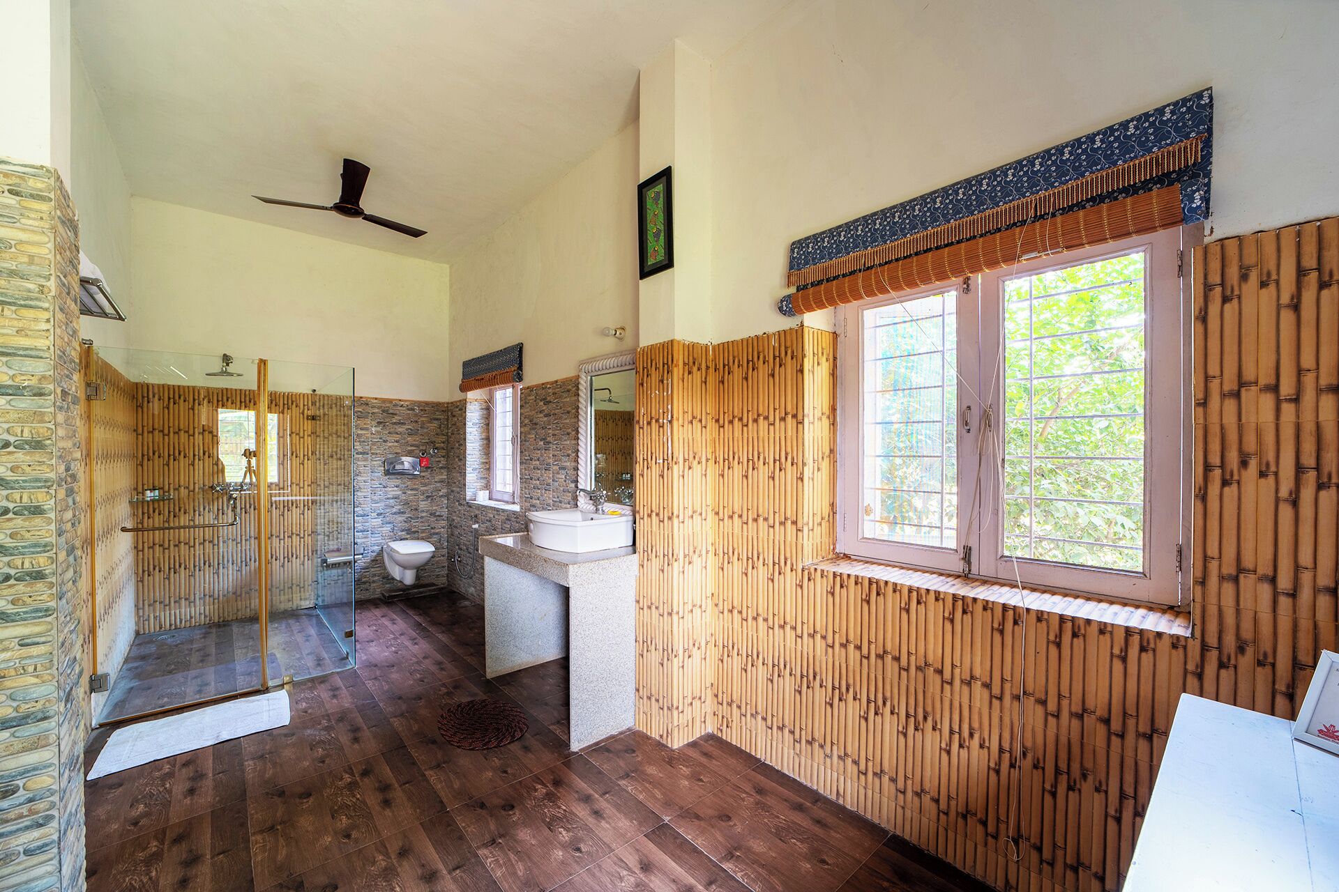 Een badkamer met een stenen muur, een douchecabine van glas, een toilet, een wastafel en een raam met bamboepaneel.