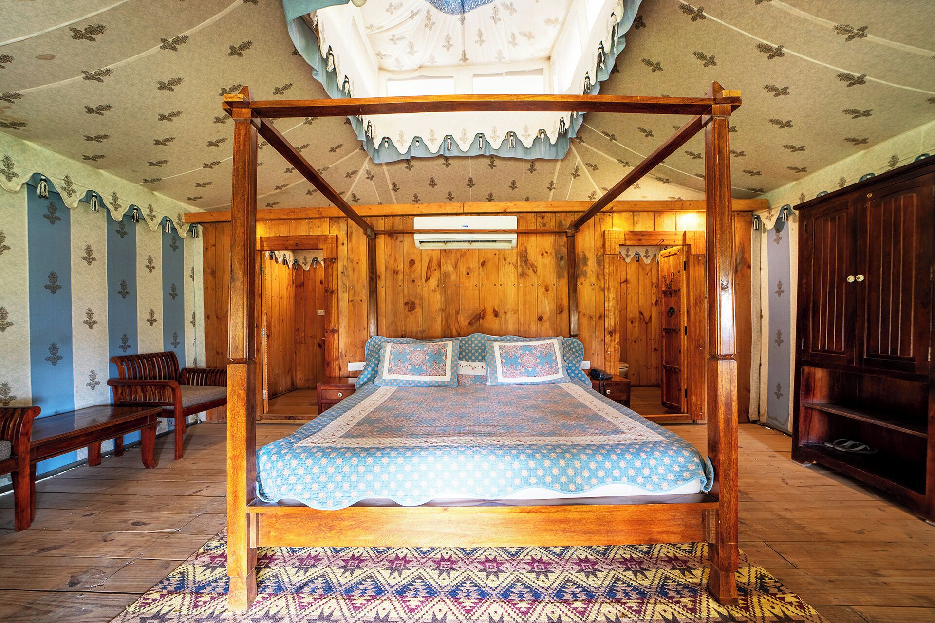 A four-poster bed with a patterned bedspread, wooden walls, and a patterned rug.