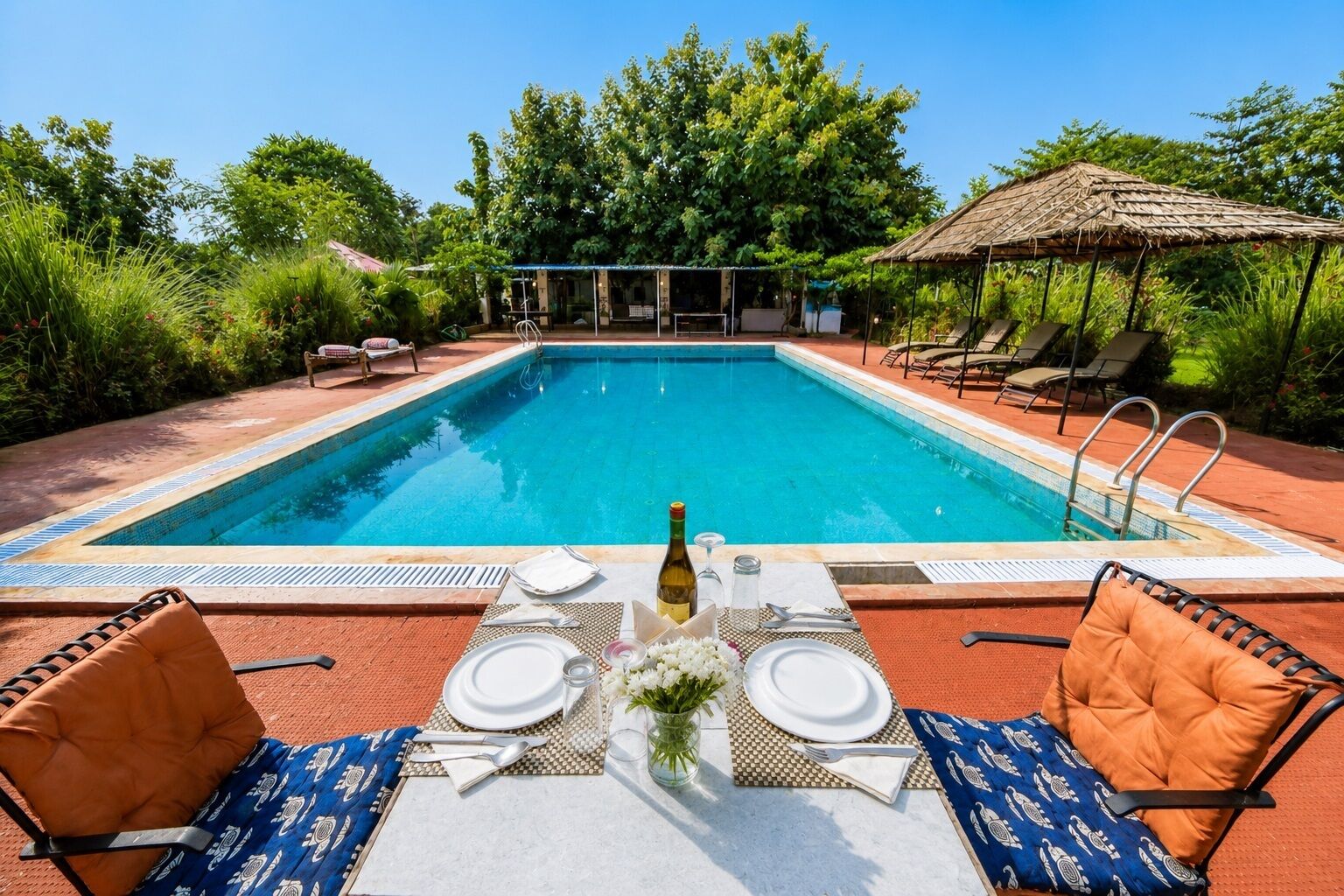 Un área de comedor junto a la piscina con una mesa preparada para dos personas, rodeada de sillones de descanso y una estructura con techo de paja.