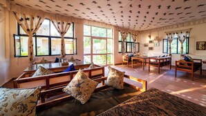 A spacious room with a high ceiling, large windows, and a variety of seating arrangements including sofas and a wooden table. - Maa Ashapura Resort (Sawai Madhopur)