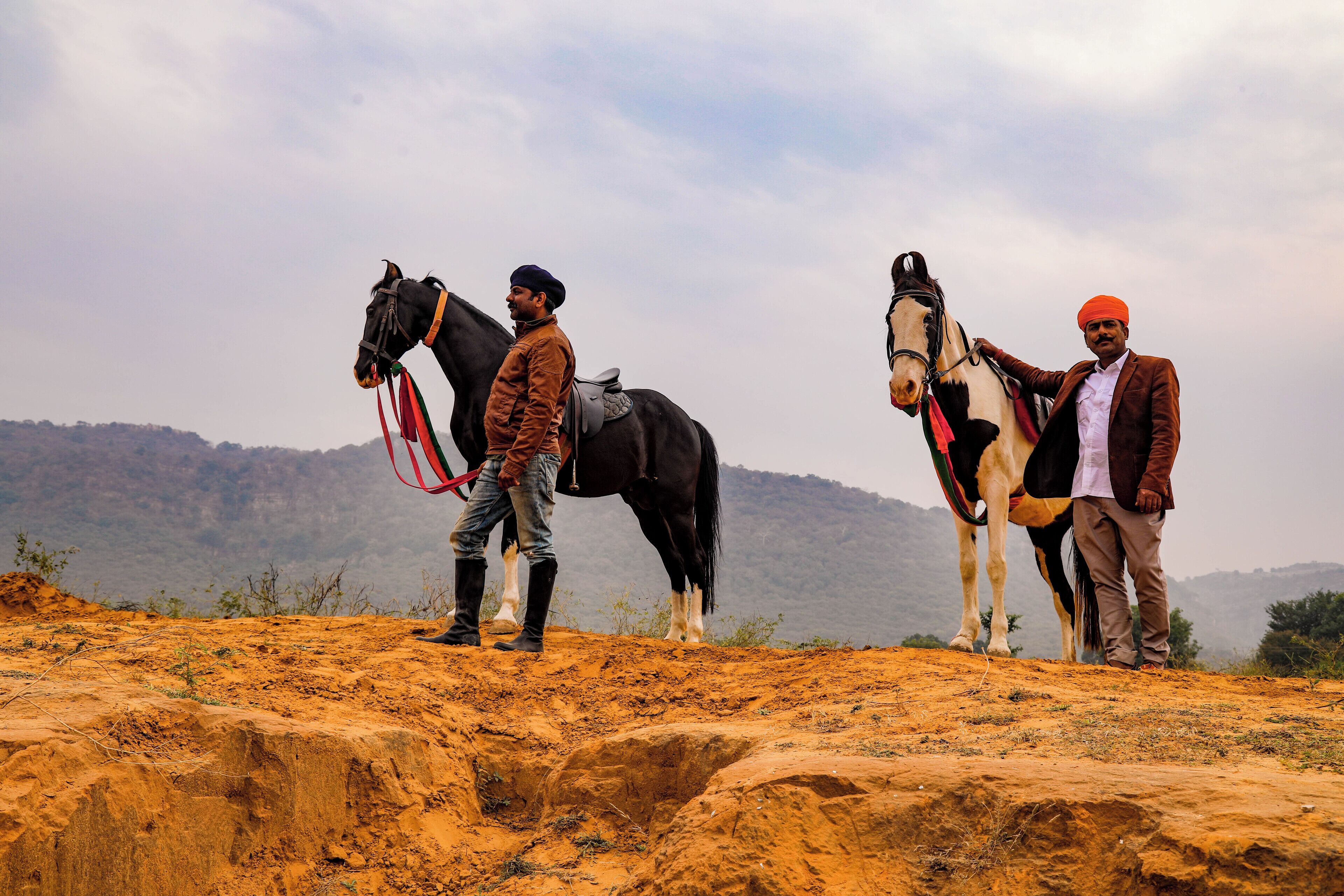 Two men with horses on a rocky terrain with a mountainous backdrop.