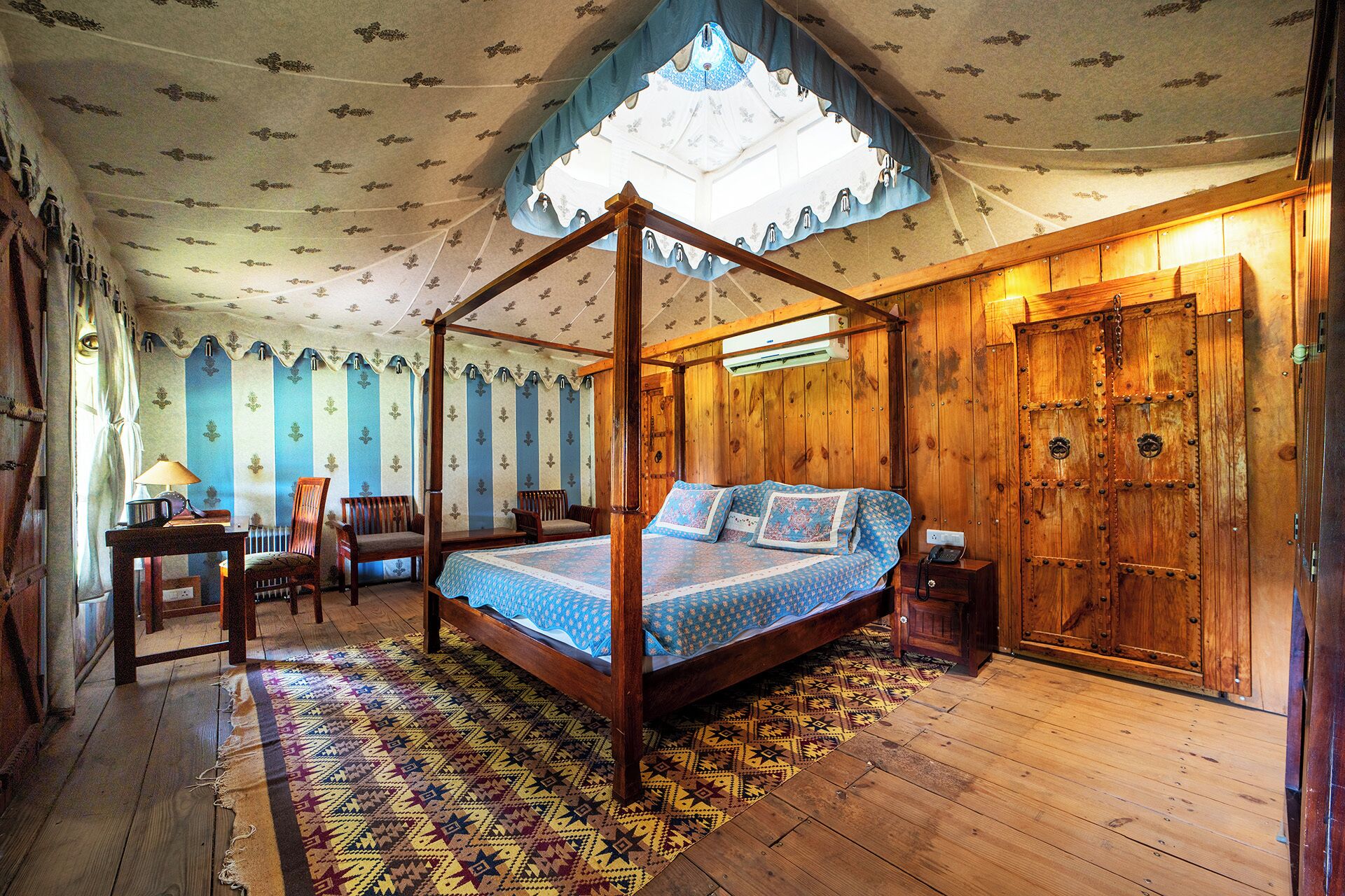 A four-poster canopy bed with a patterned bedspread, wooden floor, and a large, ornate door.