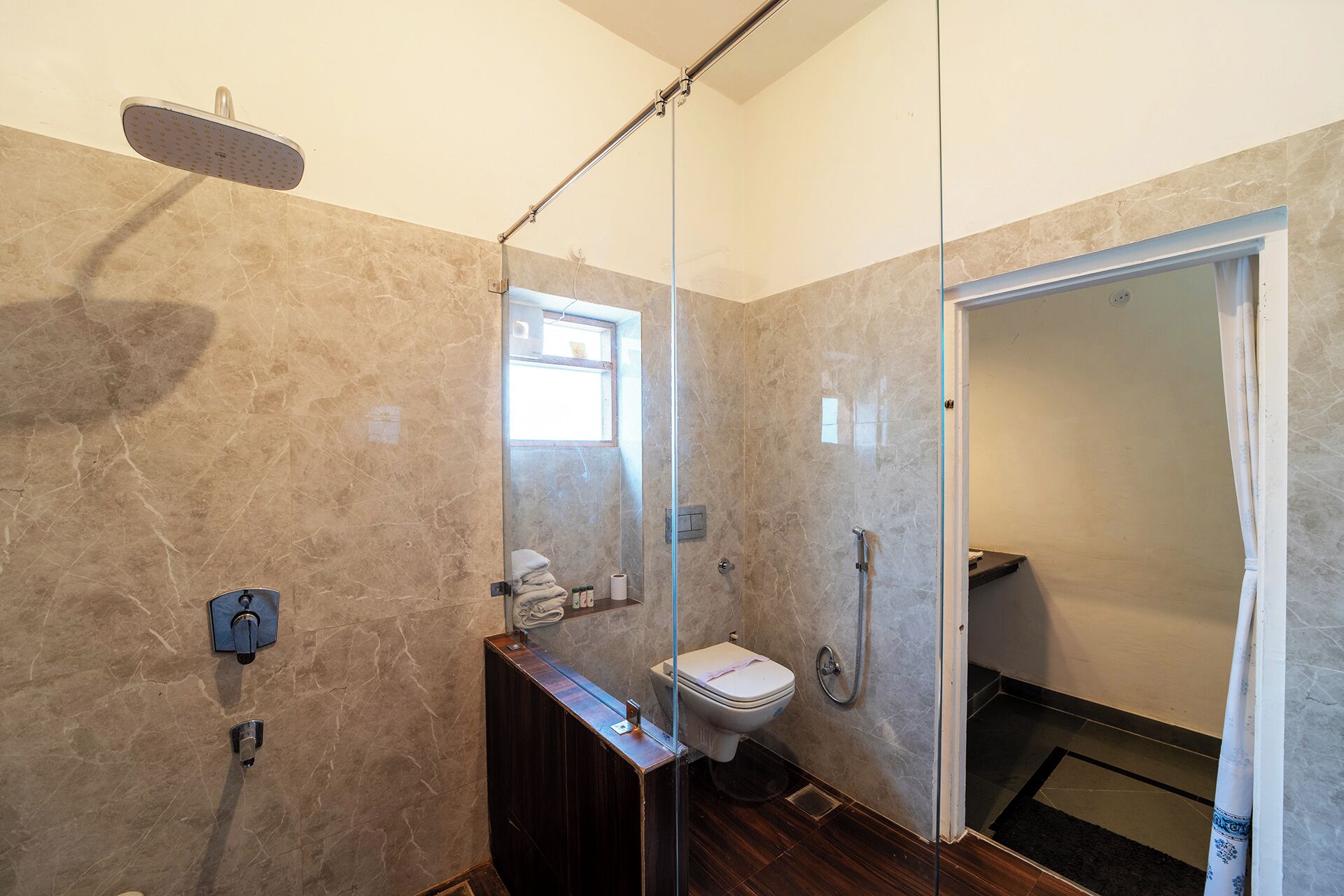 A modern bathroom with a glass shower enclosure, a wooden vanity, a toilet, and a glass door leading to another room.