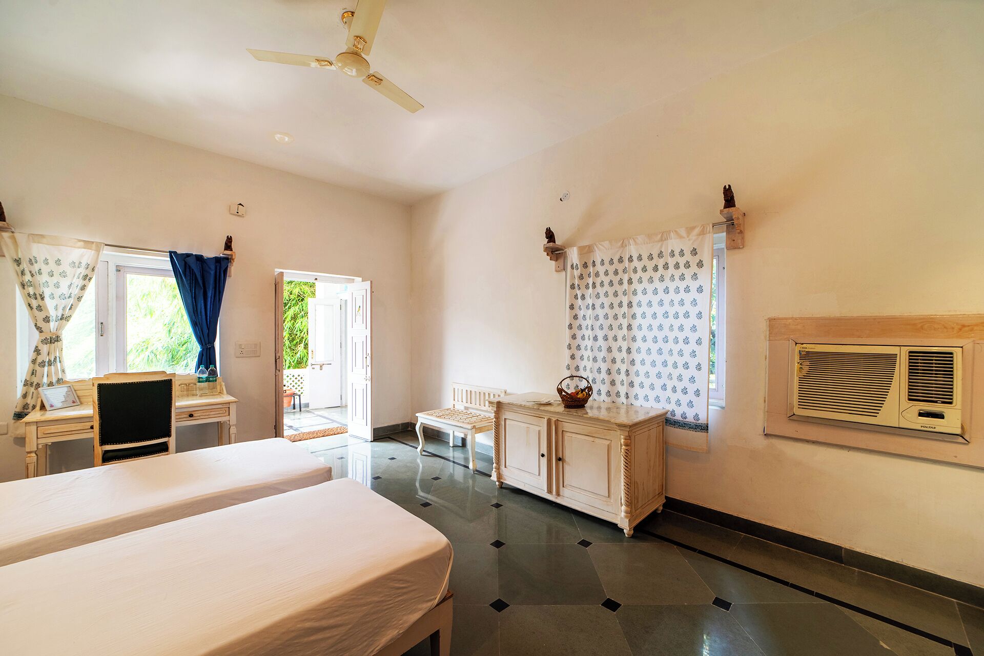 A hotel room with two beds, a desk, a chair, a wardrobe, and a ceiling fan.