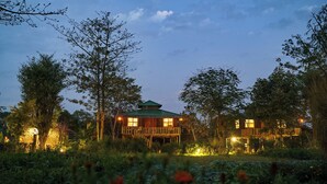 A rustic wooden house with a green roof, surrounded by trees and a garden at dusk. - Maa Ashapura Resort (Sawai Madhopur)