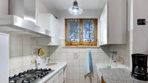 Una cocina con armarios blancos, estufa a gas, ventana con cortinas de encaje, un lavabo y una cafetera.