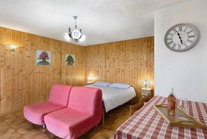 Quarto com parede de madeira, cama com colcha listrada, sofá rosa, mesa de madeira com garrafa e copo, e um relógio de parede.