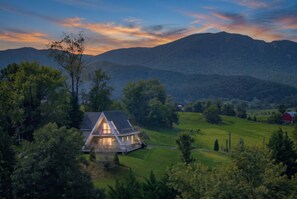 A house with a sloped roof, surrounded by a lush green landscape and mountains in the background. - Sojourners Rest: Old Rag Views, Hot Tub, Fire Pit (Etlan)