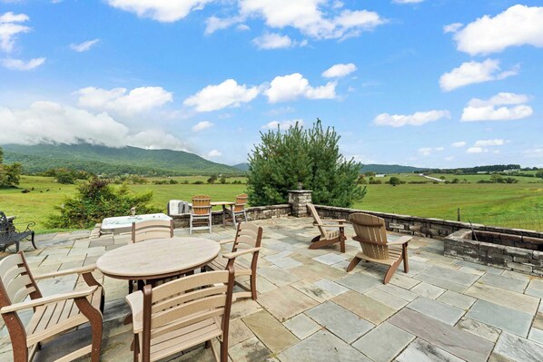 An outdoor patio with stone flooring, wooden furniture, and a view of a green landscape with hills.