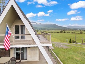 A two-story house with a balcony, a flag, and a view of a green field and mountains. - Sojourners Rest: Old Rag Views, Hot Tub, Fire Pit (Etlan)