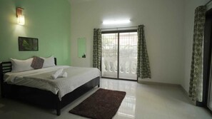 A bedroom with a bed, a nightstand, a wall-mounted light, a window with curtains, and a view of the outside. - Tokyo Villa By Staymatic Villa's (Mawal)