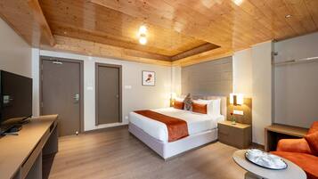 Quarto de hotel moderno com teto de madeira, cama com roupa de cama branca e detalhes em laranja, escrivaninha com televisão e uma pequena mesa com bandeja.