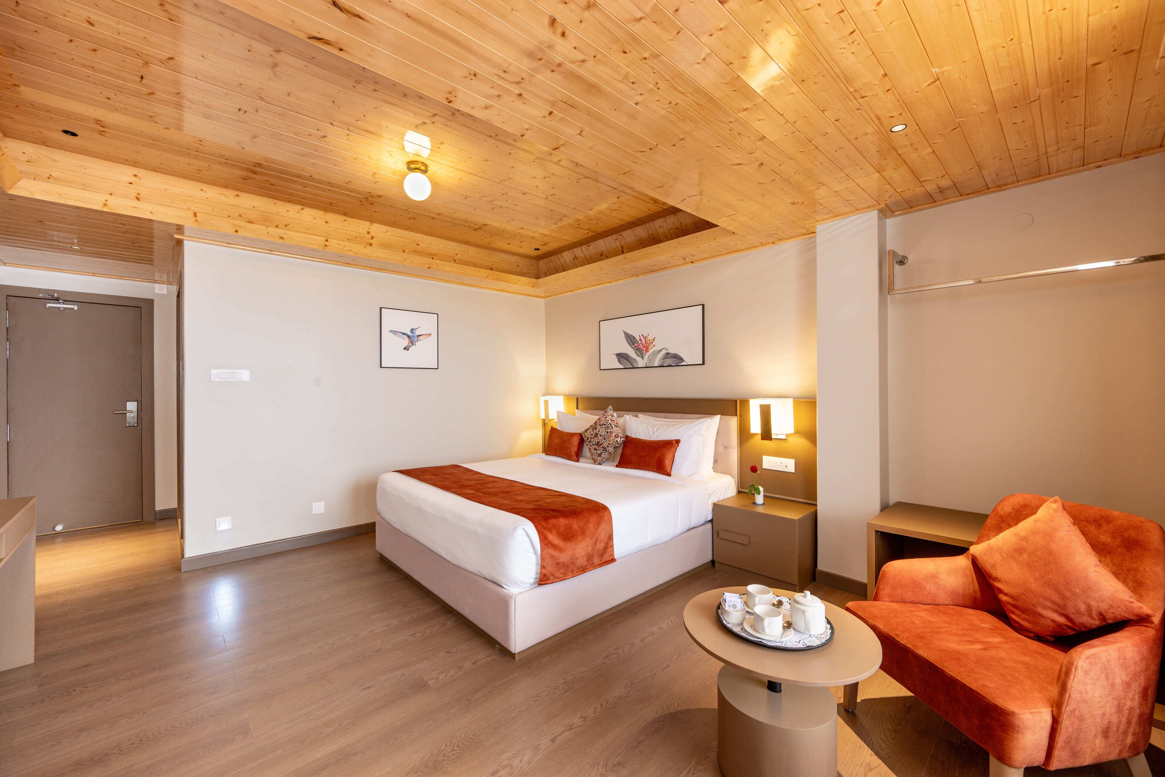 A hotel room with a wooden ceiling, a bed with a white and brown bedspread, a nightstand with a lamp, a round table with a tray of tea, and a plush orange armchair.