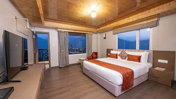 Una habitación de hotel moderna con techo de madera, una cama grande con ropa de cama blanca y naranja, un escritorio con una computadora y vistas a las montañas a través de las ventanas.