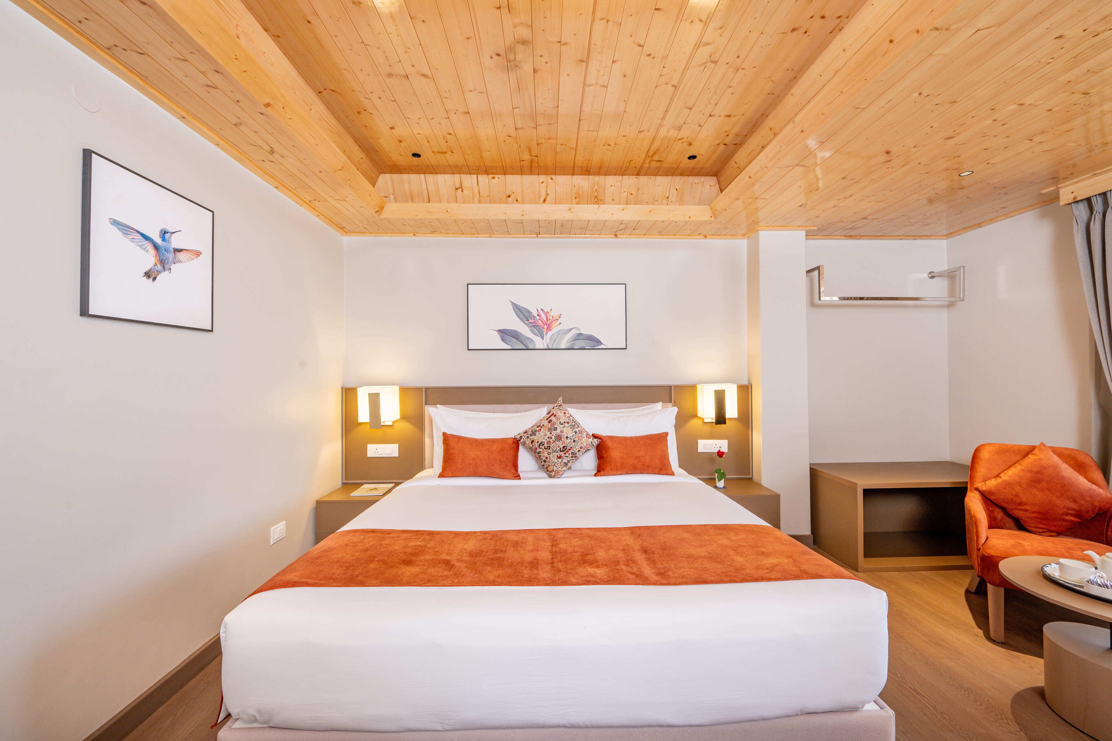 Une chambre d’hôtel avec un plafond en bois, un grand lit avec une literie orange, deux tables de chevet avec des lampes, une petite table avec une chaise et des œuvres d’art accrochées aux murs.