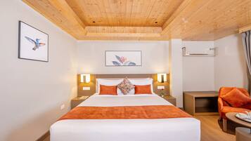 Une chambre d’hôtel avec un plafond en bois, un grand lit avec une literie orange, deux tables de chevet avec des lampes, une petite table avec une chaise et des œuvres d’art accrochées aux murs.