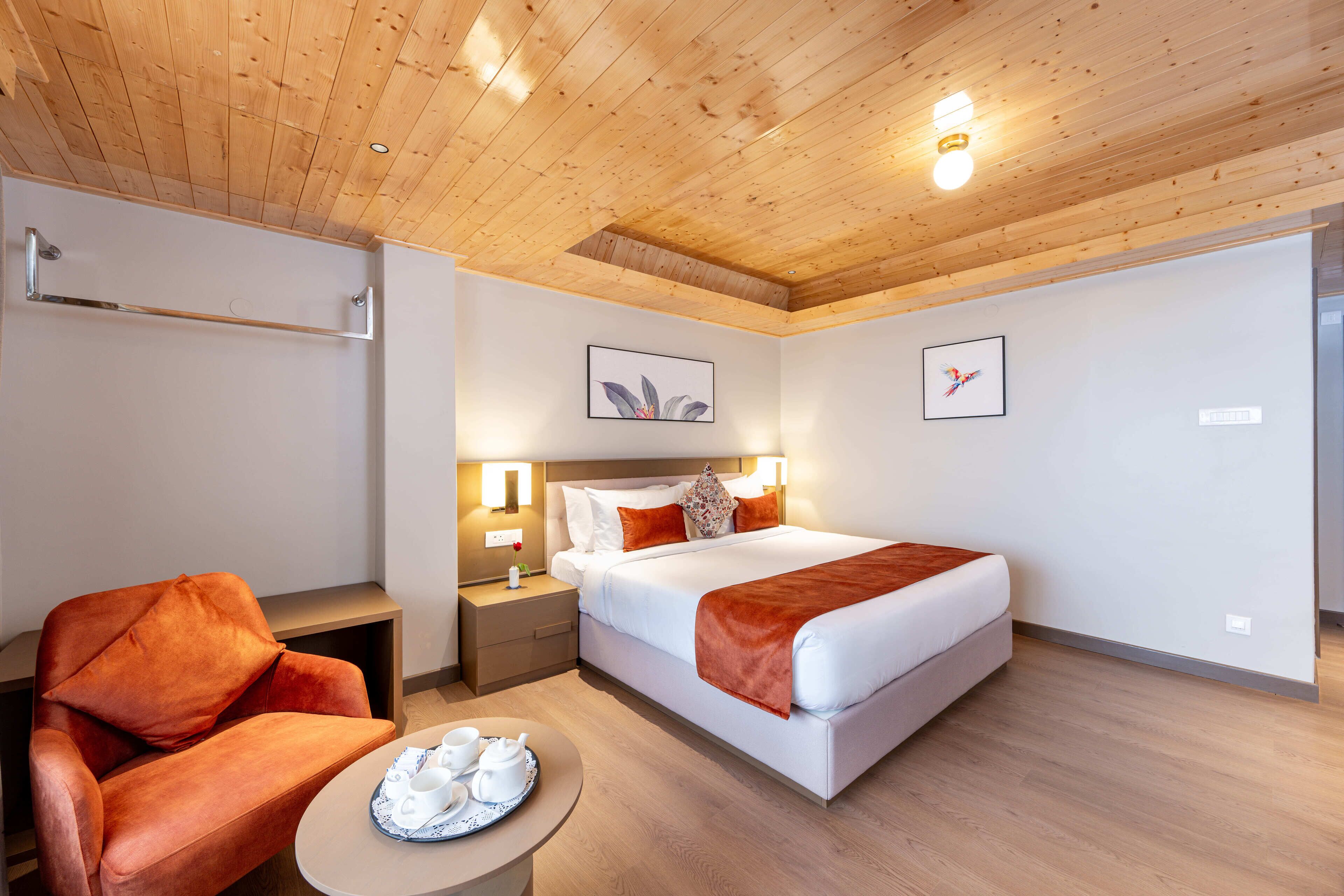 Une chambre d’hôtel avec un plafond en bois, un lit avec une literie blanche et un plaid marron, une table de chevet avec une lampe, un fauteuil rouge et une petite table sur laquelle se trouve un plateau avec des tasses et des soucoupes.