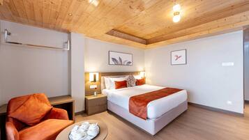 Une chambre d’hôtel avec un plafond en bois, un lit avec une literie blanche et un plaid marron, une table de chevet avec une lampe, un fauteuil rouge et une petite table sur laquelle se trouve un plateau avec des tasses et des soucoupes.