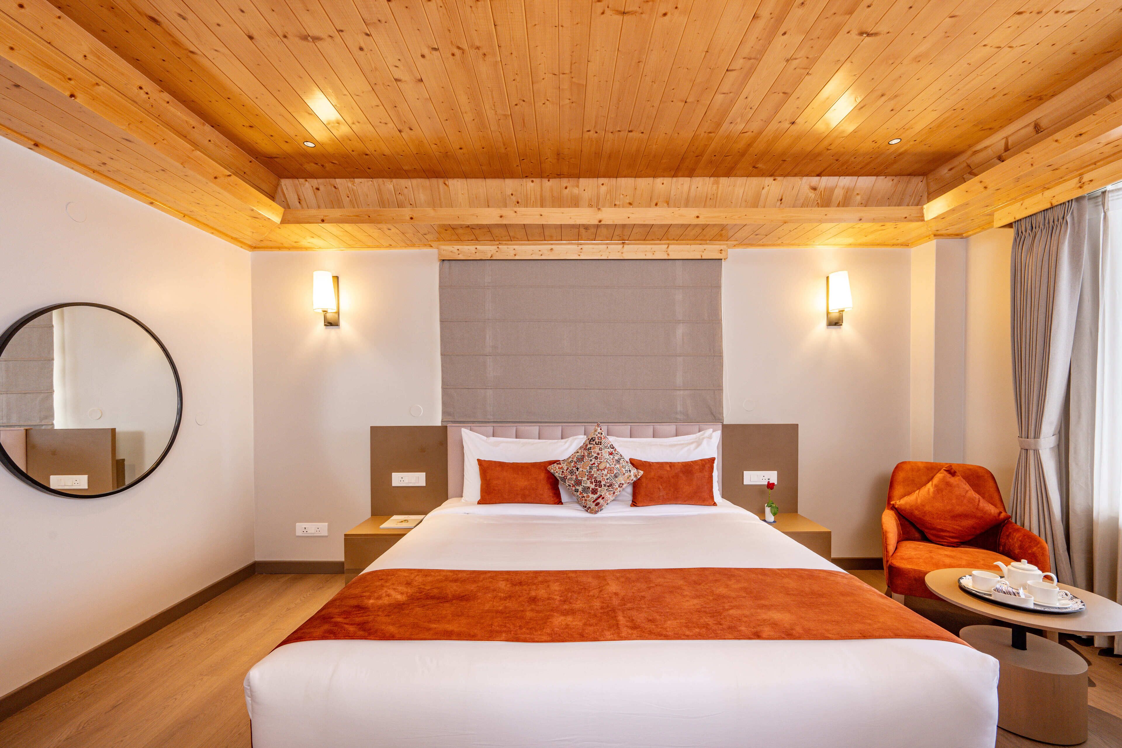 Ein Hotelzimmer mit Holzboden, einem großen Bett mit orangefarbener Bettwäsche, einem runden Spiegel, zwei Nachttischen, einem Stuhl und einem kleinen Tisch mit Tablett.