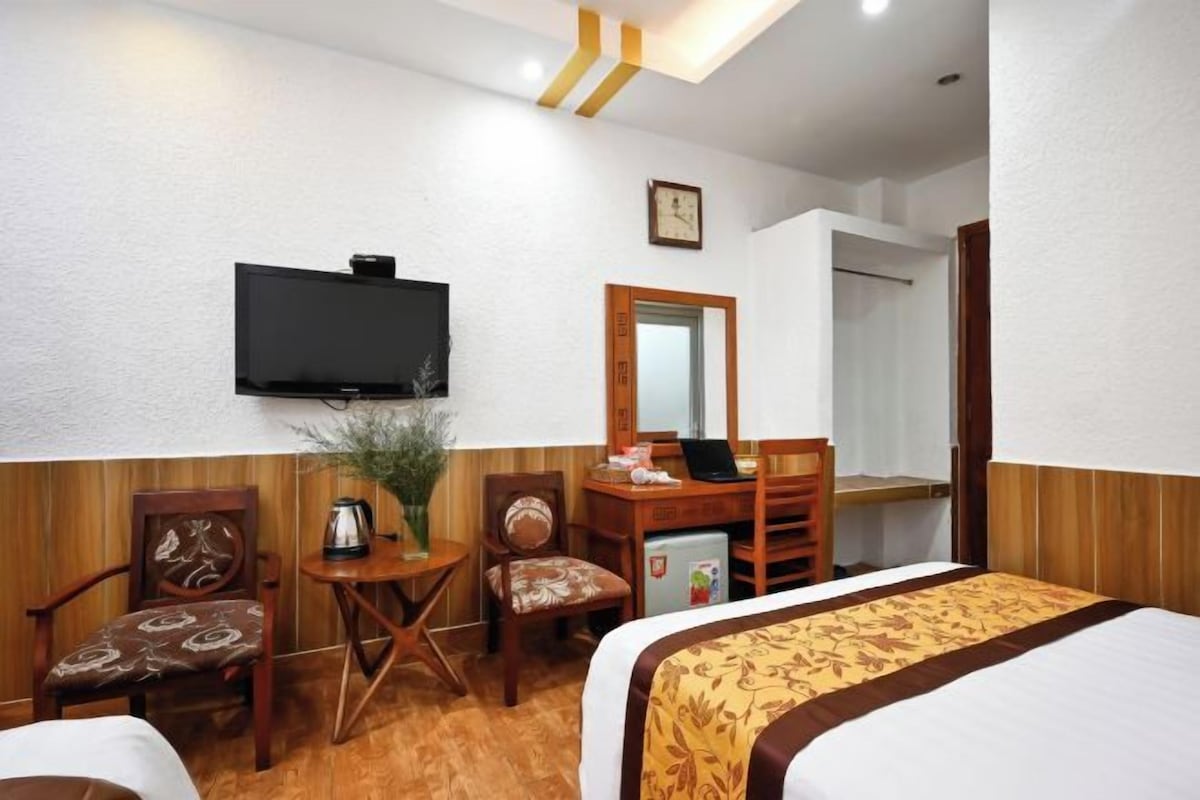 A hotel room with a bed, a desk with a computer, a chair, a small table with a plant, and a television mounted on the wall.
