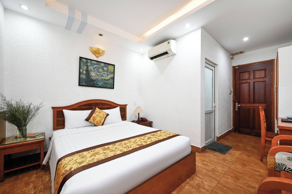Une chambre d’hôtel bien aménagée, avec un grand lit, une table de chevet avec une lampe, une petite table avec un vase, une chaise en bois et une porte.