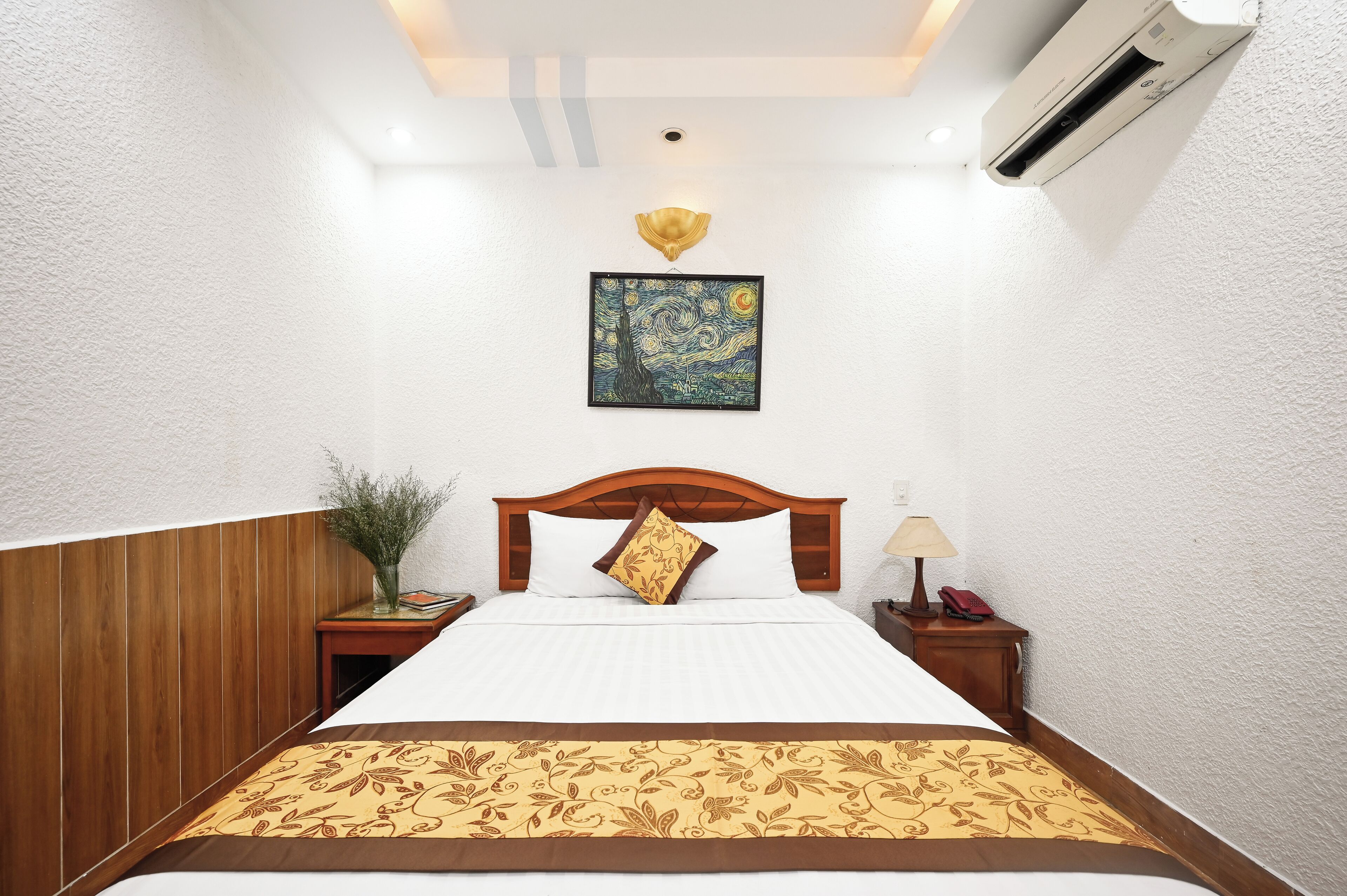 A neatly made bed with a patterned bedspread, a wooden headboard, a wall-mounted air conditioner, a framed artwork above the bed, a bedside table with a lamp and a book, and a small potted plant.