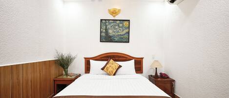 A neatly made bed with a patterned bedspread, a wooden headboard, a wall-mounted air conditioner, a framed artwork above the bed, a bedside table with a lamp and a book, and a small potted plant.