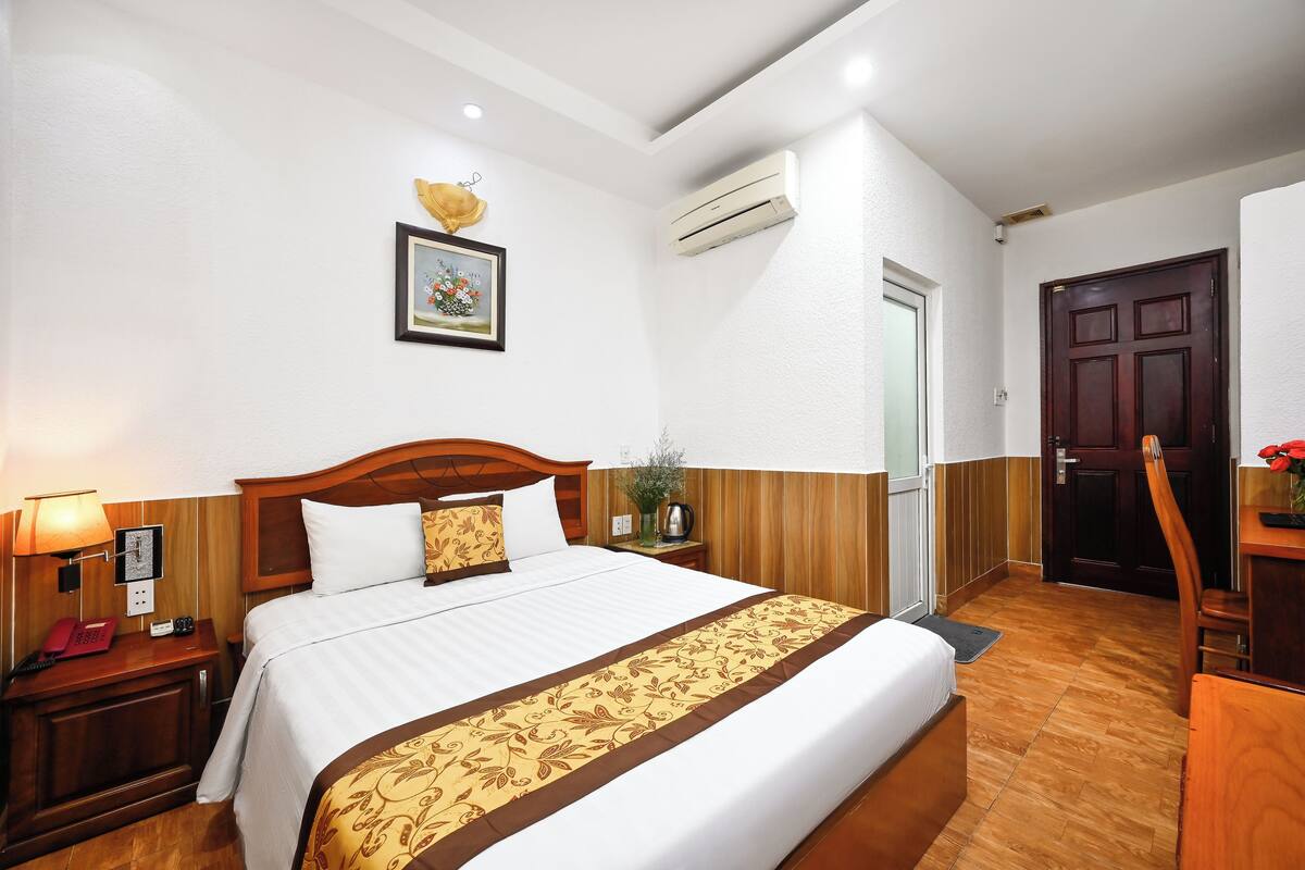 Une chambre d’hôtel avec un grand lit, une tête de lit en bois, des tables de chevet avec des lampes, un tableau encadré au mur, un appareil de climatisation et une porte en bois.