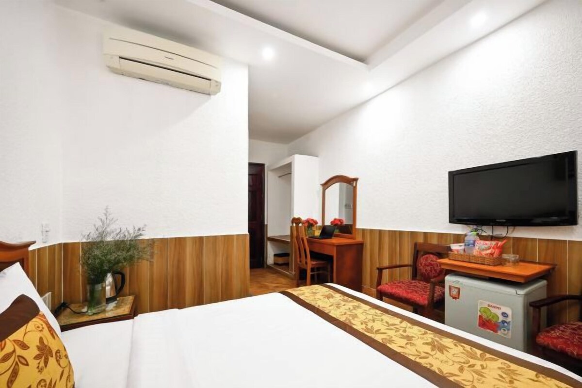 A hotel room with a bed, a desk with a chair, a television mounted on the wall, and an air conditioning unit.