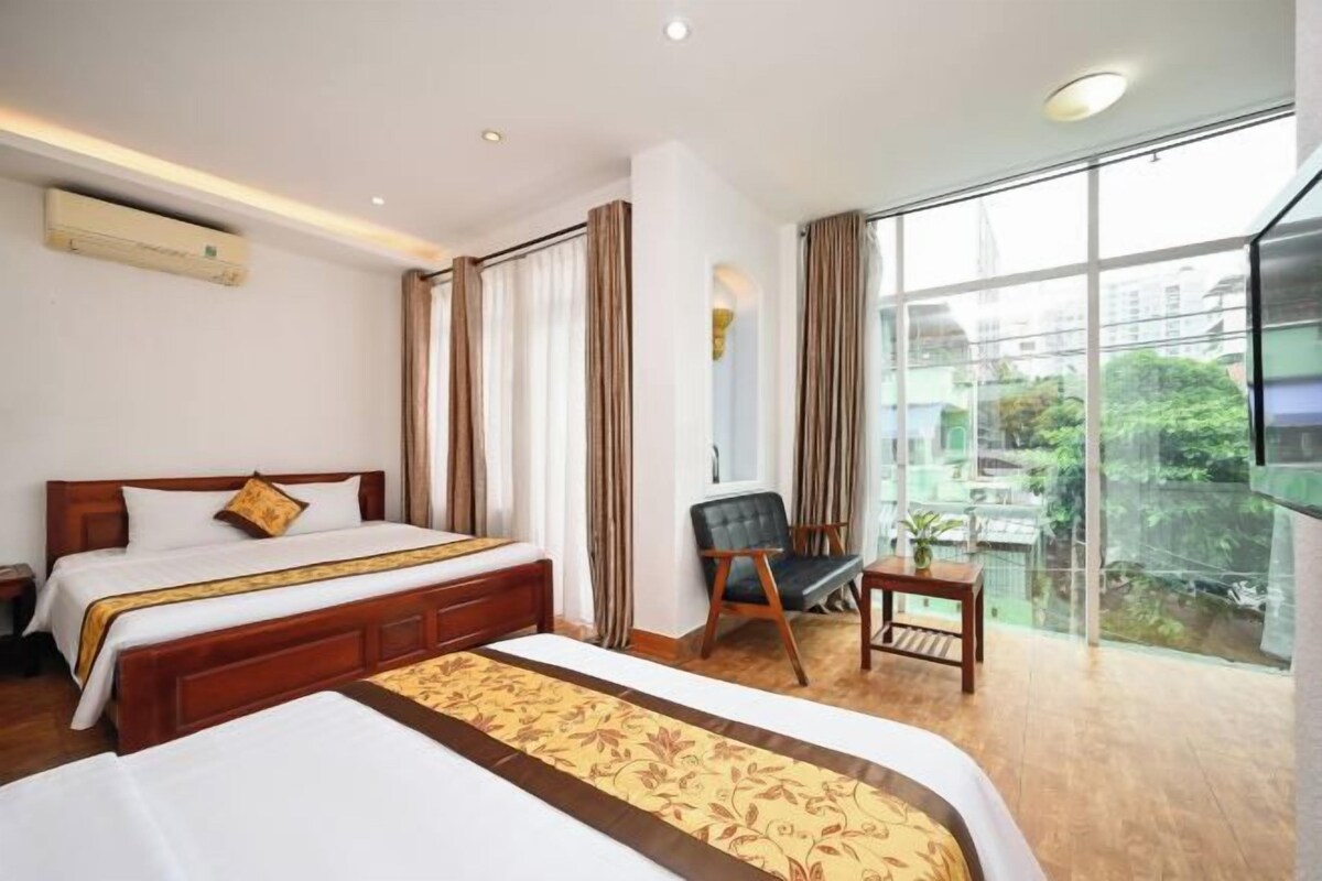 A hotel room with a large bed, a small desk, a chair, and a view of greenery through large windows.