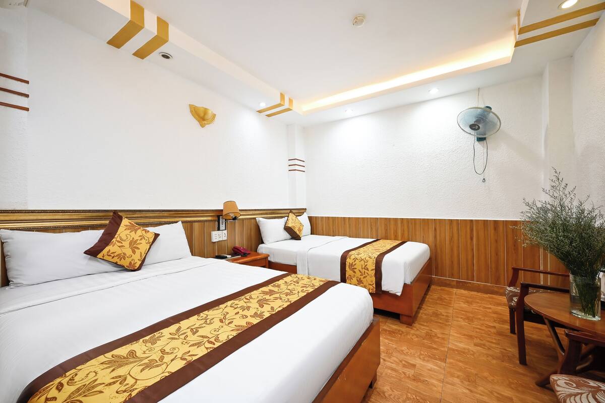 Une chambre d’hôtel avec deux lits, des têtes de lit en bois, du linge de lit blanc, un plancher en bois, une table avec des chaises et un ventilateur fixé au mur.