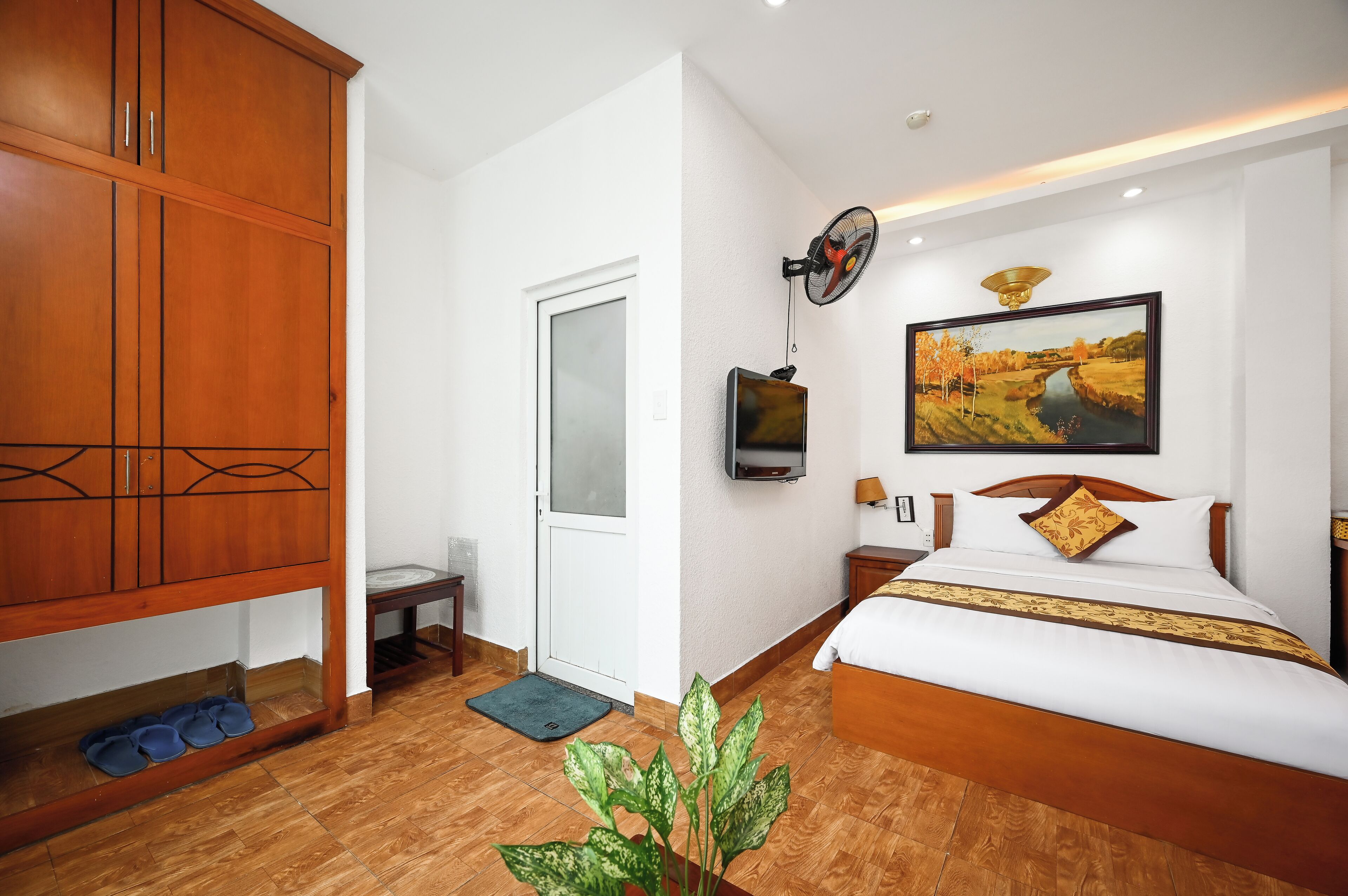 A bedroom with a wooden wardrobe, a bed with a white and gold bedspread, a painting on the wall, a ceiling fan, and a television.
