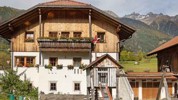 A wooden chalet with a balcony, a flag, and a mountain backdrop. - Apartment 'Inneroswalderhof  3' with Mountain View, Shared Garden and Wi-Fi (Anterselva di Sotto)