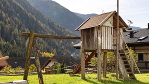 A wooden playhouse with a slide and a ladder, surrounded by a grassy area and houses, with mountains in the background. - Apartment 'Inneroswalderhof  3' with Mountain View, Shared Garden and Wi-Fi (Anterselva di Sotto)
