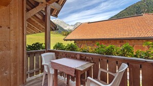 Une terrasse en bois avec une table et des chaises, d’où l’on peut admirer une montagne et un bâtiment dont le toit est recouvert de tuiles.