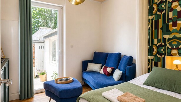 Een slaapkamer met een bed, een bank, een houten vloer, een plafondlamp en uitzicht door een deur.