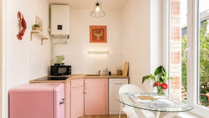 Een kleine keuken met een roze koelkast, een magnetron en een spoelbak. Er is een glazen tafel met stoelen, een potplant en een raam met uitzicht op groen.