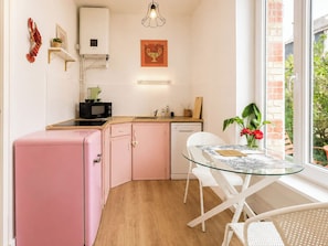 Cocina compacta con refrigerador rosa, microondas y lavamanos. Hay una mesa de vidrio con sillas, una planta en maceta y una ventana con vista a follaje.