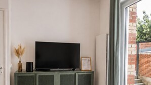 Een moderne woonkamer met een flatscreen-tv, een groen kastje, een blauwe poef en een groot raam met uitzicht op een bakstenen muur en groen.